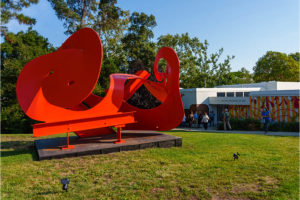 A large-scale steel sculpture painted bright red. Loops of metal remiscent of a mobius strip. Museum beyond.