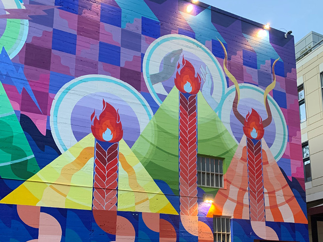 Close up image of Maria Molteni's mural in downtown SLO. Quilt-like patterns of semi-circles falling down before a minimalistic water background. Geometric mountains with braids of lava, symbolizing the seven sisters of SLO. Seven stars in quilt-like patterns representing the Pleiades are above the mountain peaks.
