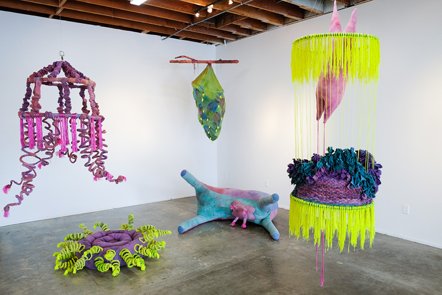 Ensemble of soft sculptures by Marrin Lee. A cement-floor gallery with soft sculptures in bright colors hanging from the ceiling. 2 soft sculptures, 1 like a cow and the other like a sea anemone, lie on the floor.