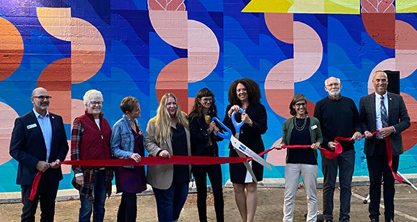 9 people cut a ceremonial ribbon for Maria Molteni's mural in downtown SLO, "Seven Sisters"