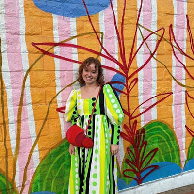 young woman in green/white/black stripped dress and red bag stands in front of a colorful mural
