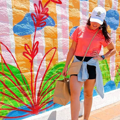 Young woman in white Cal Poly SLO hat and orange t-shirt stands in front of a colorful mural