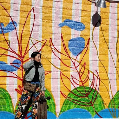 a young woman in a gray jacket, floral pants, and holding a skateboard stands in front of a colorful mural