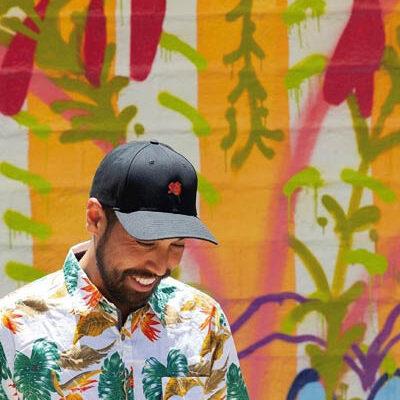 a young man in a black hat and tropical-pattern shirt looks down and laughs, a colorful mural behind him