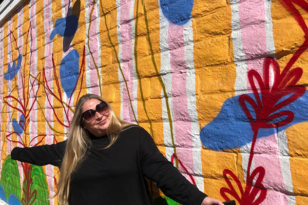 SLOMA executive director, Leann Standish, wearing a black sweater and sunglasses, stands in front of a colorful mural