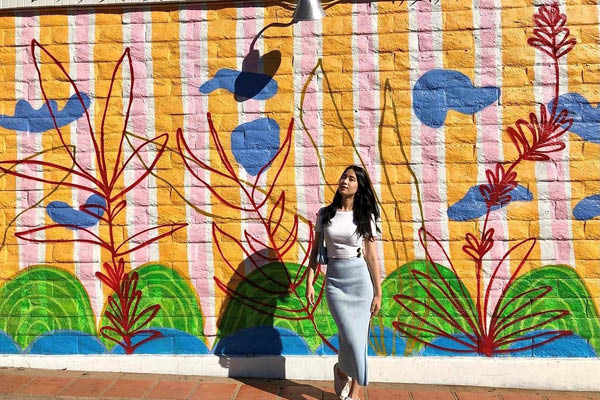 a young woman in a white shirt and light blue long skirt stands in front of a colorful mural