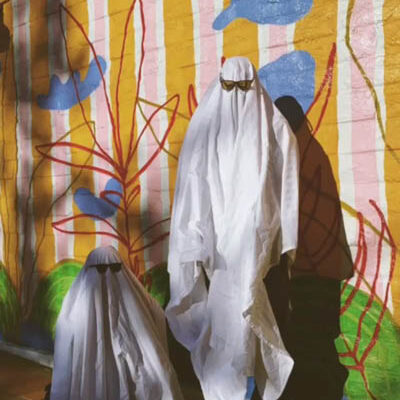 two people dressed as ghosts wear sunglasses, one standing and one kneeling in front of a colorful mural