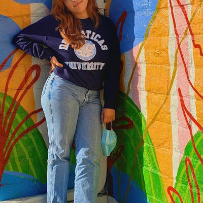 young woman in a college sweatshirt and jeans leans up against a colorful mural