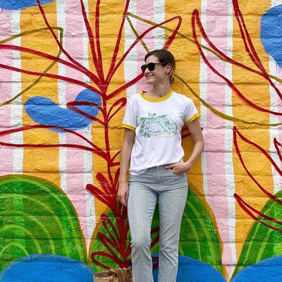 young woman in t-shirt and cropped jeans leans against a colorful mural