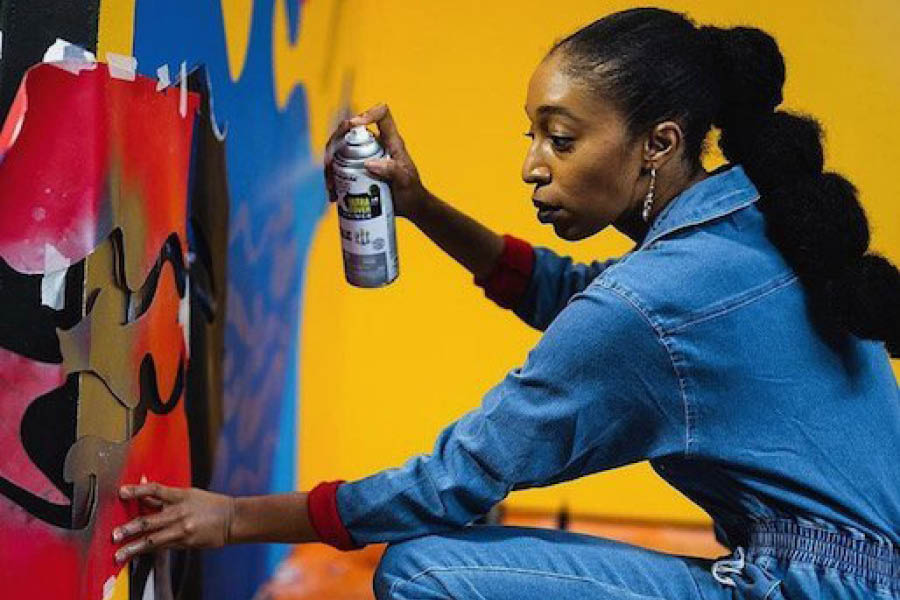 A woman in blue overalls uses spray paint and a large stencil to work on a mural