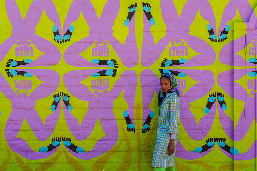 A portion of Erin LeAnn Mitchell's mural, "Calafia Was Here," with the artist depicted. A black woman wearing a light blue long coat and handkerchief on her head stands before a mural depicting purple 4-legged figures on a lime green background.