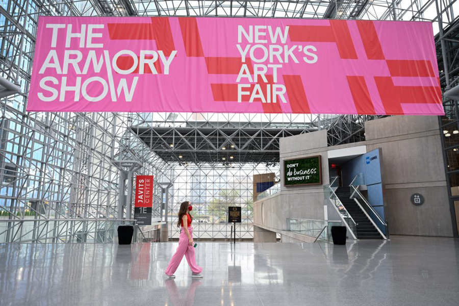 The Armory Show: New York's Art Fair. A bright pink banner hangs from an industrial building's ceiling, as a young woman in pink pants walks below
