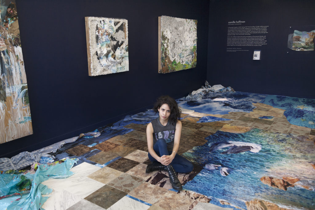 Camille Hoffman sitting cross-legged on the floor of one of her exhibitions. The walls are painted dark blue and the floor is covered with colorful vinyl squares