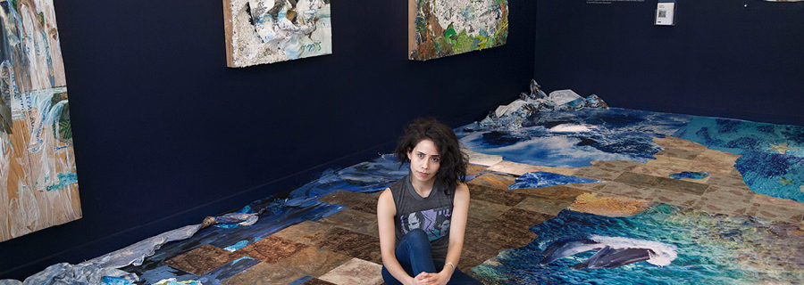Artist Camille Hoffman, a woman with dark hair wearing a sleeveless tee, sits cross-legged among her artworks