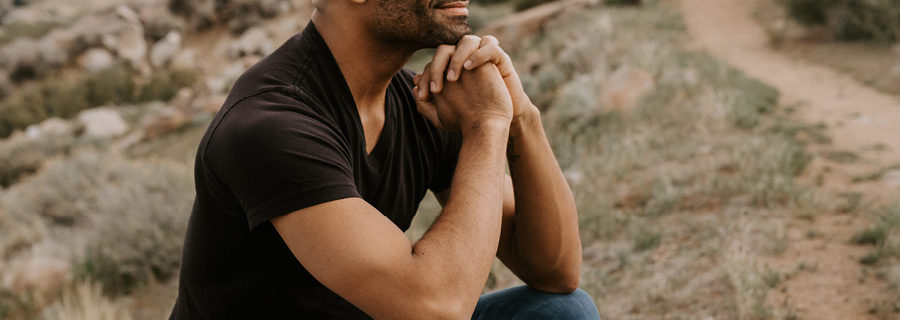 Yoga and mindfulness instructor Jevon Rowden, a Black man wearing a black v-neck t-shirt and blue jeans, sits on a hillside with his hands under his chin