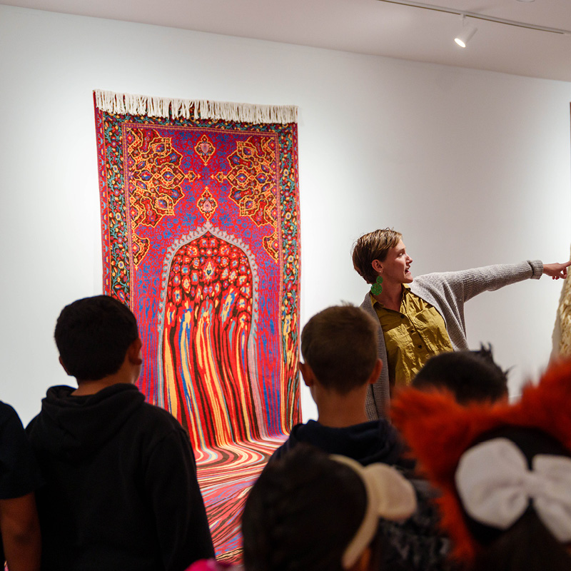 A group of 2nd graders, shown from behind, listen to SLOMA's Chief Curator on a school tour of Faig Ahmed's exhibition