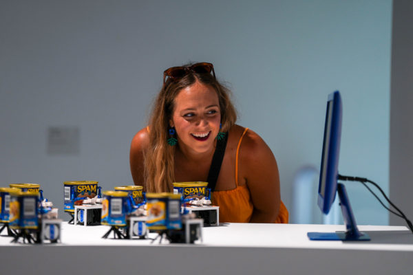 A woman in a yellow top and sunglasses smiles next to "Spambots," an artwork by Neil Mendoza utilizing little cans of SPAM, each with four keyboard keys. The bots collectively type pork-powered gibberish onto a computer screen