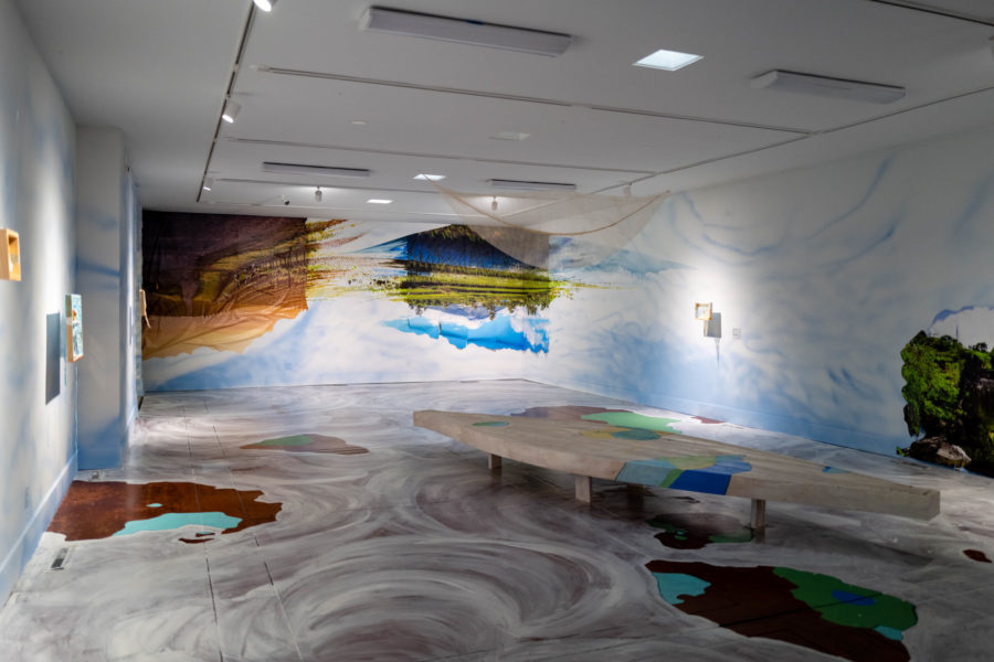 View of the west wall of the Gray Wing, showing Camille Hoffman's artwork in "See and Missed." Images of Philippine farm lands and California vineyards hang upside down in long vinyl strips. The bottoms (skies) are torn and folded and the images seemingly blend together through paint. The floor is painted to resemble islands in a swirling white sea.