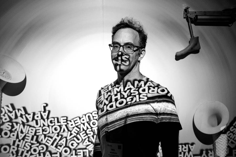 Head shot of artist Neil Mendoza. A man wearing glasses stands facing the camera at a slight angle, his projected artwork of jumbled words partially obscures his face