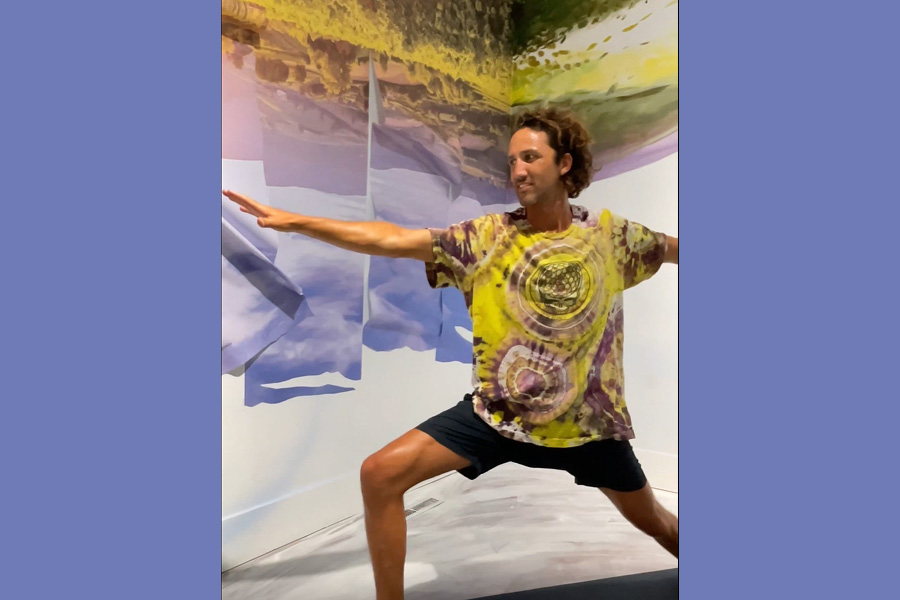 Yoga instructor Charley Newel. A man with brown curly hair, wearing a yellow and pink tie-dyed shirt, forms the warrior pose in SLOMA's Gray Wing. Swirling purples and greens on the wall behind him.