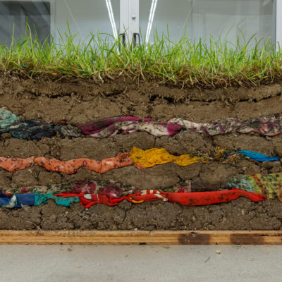 An art installation by Chilean-American artist Minga Opazo. A cross section of grass and soil is interwoven with layers of textile waste