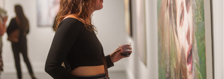 A young woman in a black top holds a glass of wine as she looks at an artwork depicting a woman in water. "GATHERING" is typed at the bottom left corner