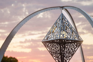 "The Greys In Between" by Anila Agha. A three-legged arch supports a hanging metallic diamond sculpture decorated with intricate scrollwork and an interior globe light to cast shadows. A beautiful sunset colors the sky beyond.