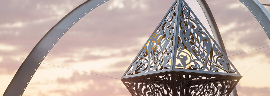 "The Greys In Between" by Anila Quayyum Agha . A three-legged arch supports a hanging metallic diamond sculpture decorated with intricate scrollwork and an interior globe light to cast shadows. A beautiful sunset colors the sky beyond.