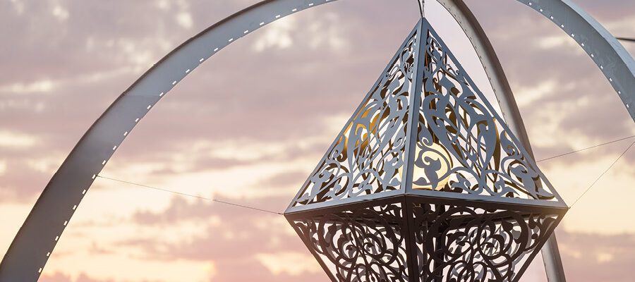 The Greys In Between by Anila Quayyum Agha "The Greys In Between" by Anila Quayyum Agha . A three-legged arch supports a hanging metallic diamond sculpture decorated with intricate scrollwork and an interior globe light to cast shadows. A beautiful sunset colors the sky beyond.