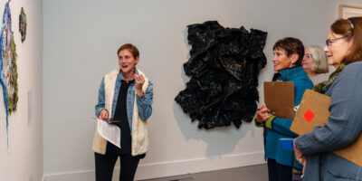 SLOMA's curator leads a docent training event. A young woman with short blonde hair and a white sweater refers to a painting while three other women holding clipboards listen and take notes for future gallery tours