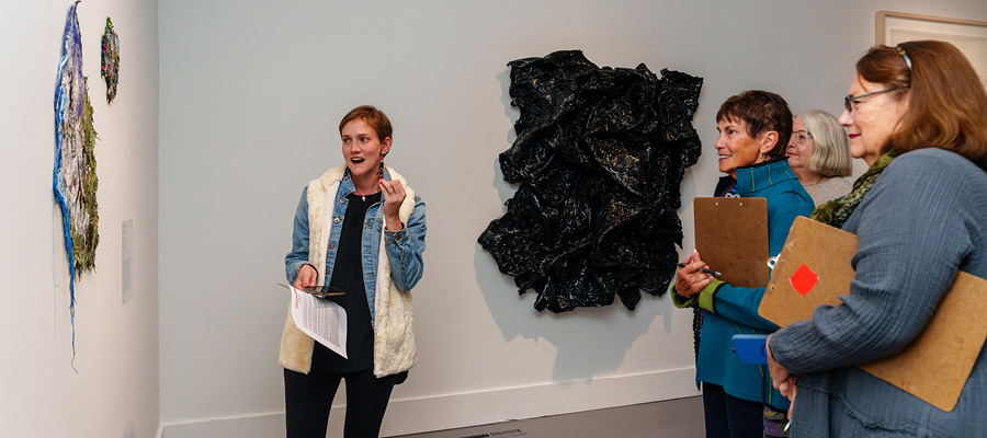 SLOMA's curator leads a docent training event. A young woman with short blonde hair and a white sweater refers to a painting while three other women holding clipboards listen and take notes for future gallery tours