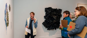 SLOMA's curator leads a docent training event. A young woman with short blonde hair and a white sweater refers to a painting while three other women holding clipboards listen and take notes for future gallery tours