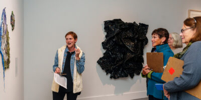 docents banner2 SLOMA's curator leads a docent training event. A young woman with short blonde hair and a white sweater refers to a painting while three other women holding clipboards listen and take notes for future gallery tours