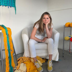 Minga Opazo headshot. A woman with long blonde hair, wearing a white t shirt and white pants, sits on a white armchair surrounded by colorful textile art