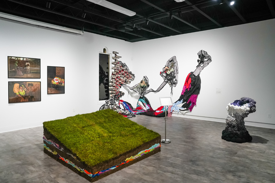 An exhibition shot of "Dirty Laundry" at SLOMA. A slanted platform is covered with grass, the sides revealing layers of soil and discarded clothing. A purple, white, and gray sculpture appears like bubbles rising from the floor. A photo collage of textile workers in India.