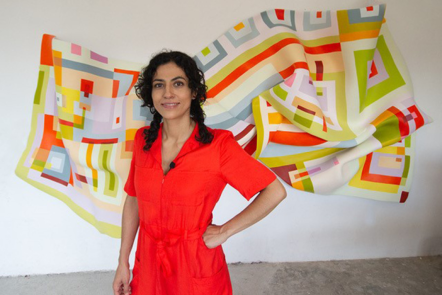 Artist Marela Zacarías, a woman with braided brown hair, wearing an orange jumpsuit stands in front of a large sculpture with folds and colorful geometric patterns