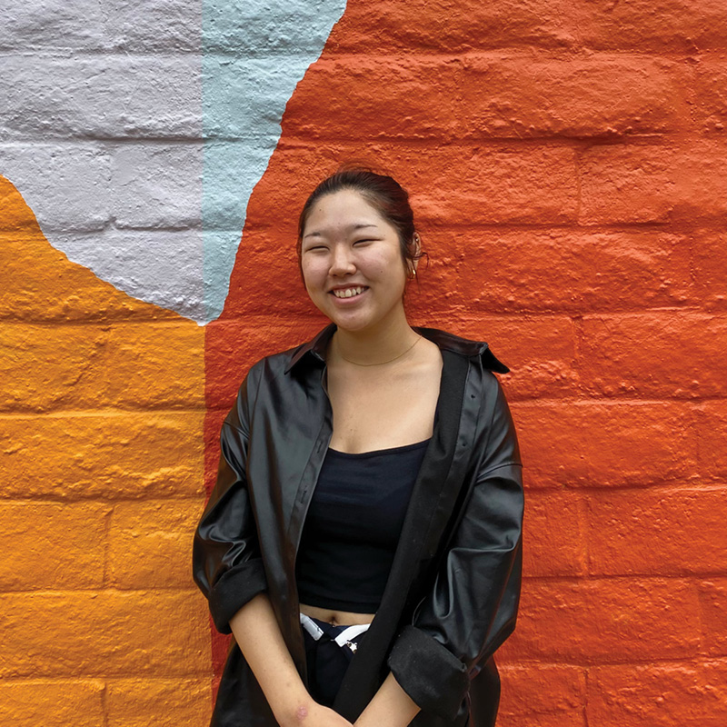Photo of Hailey Choi, SLOMA's summer 2023 intern. A young woman in a black button down shirt, black cami, and black pants smiles in front of a mural of hills