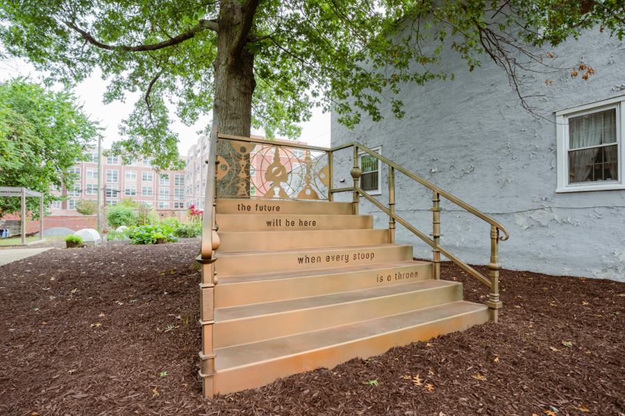 A gold-colored staircase leads to a tree. Engraved on some of the stair risers: "The future will be here when every stoop is a throne."