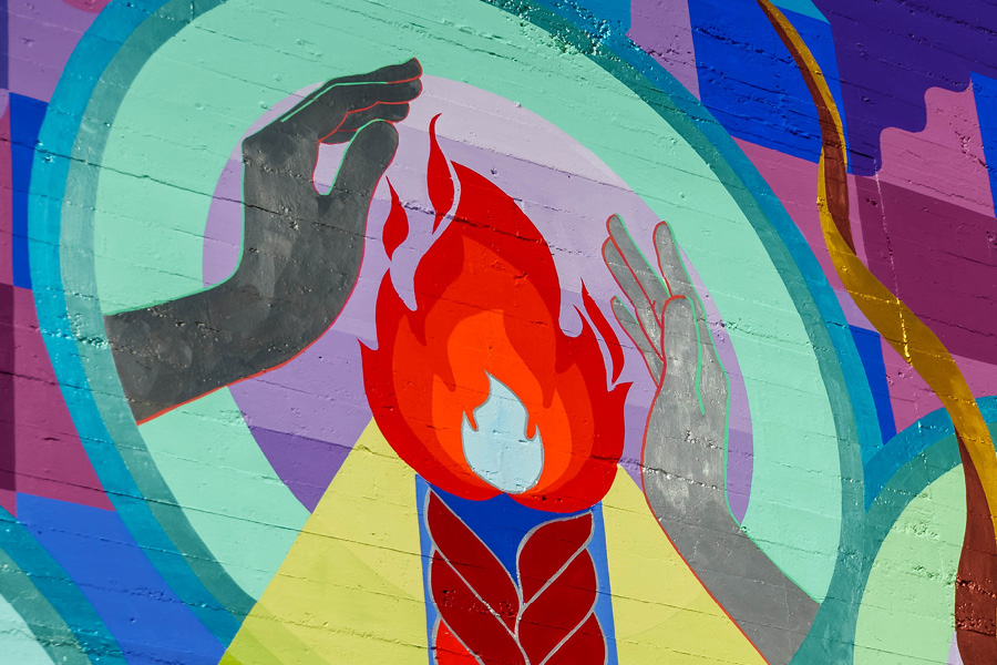 Close up image of Maria Molteni's mural in downtown SLO. Quilt-like patterns of semi-circles falling down before a minimalistic water background. Silver hands warm themselves by the fire of the mountain peak. Geometric mountains with braids of lava, symbolizing the seven sisters of SLO.