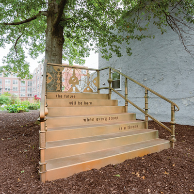 A gold-colored staircase leads to a tree. Engraved on some of the stair risers: "The future will be here when every stoop is a throne."