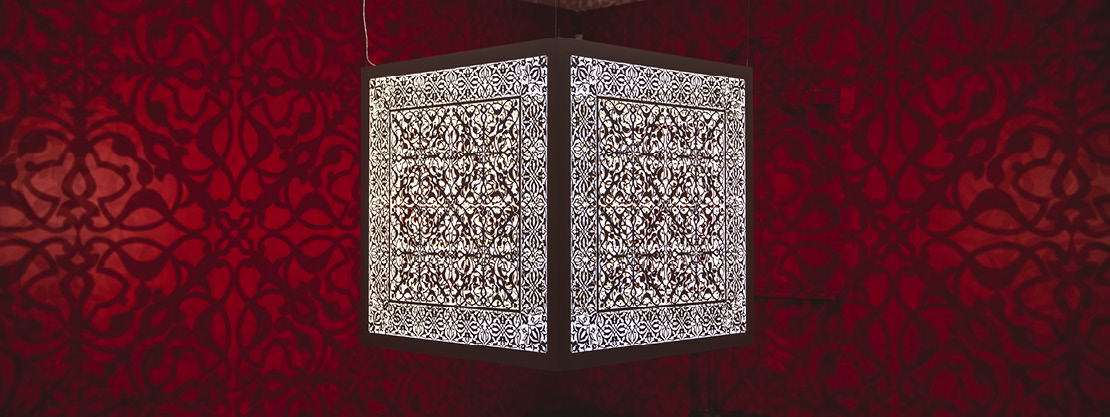 A cube of steel hangs from the ceiling, its surface delicately etched with curves and floral patterns that cast shadows from a single lightbulb within