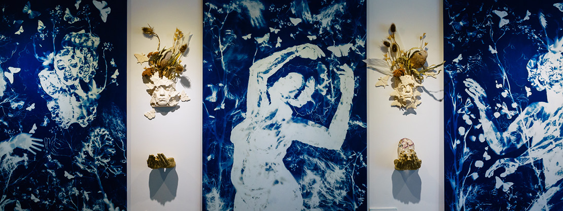 Three large cyanotype paintings of figures and flowers, with butterfly faces and hands in between the canvases