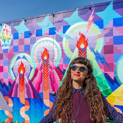 Close up image of Maria Molteni's mural in downtown SLO. Quilt-like patterns of semi-circles falling down before a minimalistic water background. Geometric mountains with braids of lava, symbolizing the seven sisters of SLO. Seven stars in quilt-like patterns representing the Pleiades are above the mountain peaks. The artist, wearing a purple polka dot blouse and sunglasses, stands before the mural.