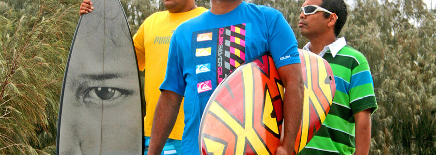 Photograph of three men on a beach wearing bright t-shirts, sunglasses, and carrying surfboards