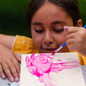 A young girl in a yellow shirt uses pink paint on a canvas