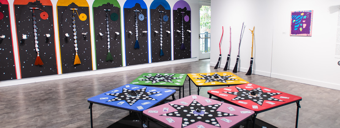 Panorama of an exhibition installation showing 5 card tables in the middle of a room with witchy brooms and celestial paintings on the walls