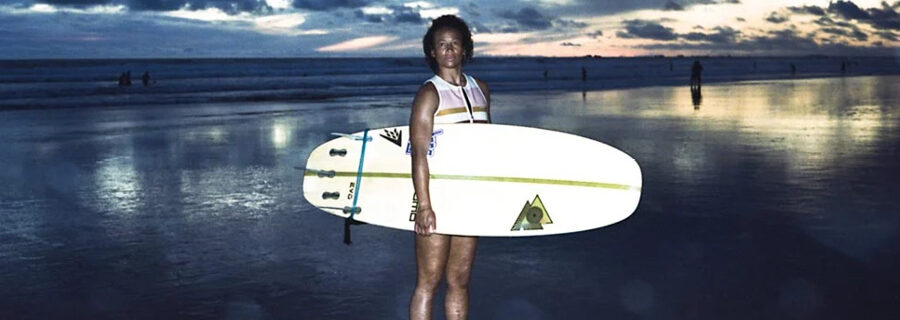 Photo by Gabriella Angotti-Jones. A young Black woman holds her surfboard, standing on the beach at sunset