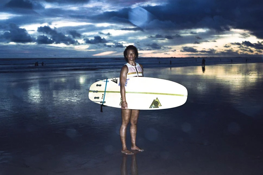 Photo by Gabriella Angotti-Jones. A young Black woman holds her surfboard, standing on the beach at sunset