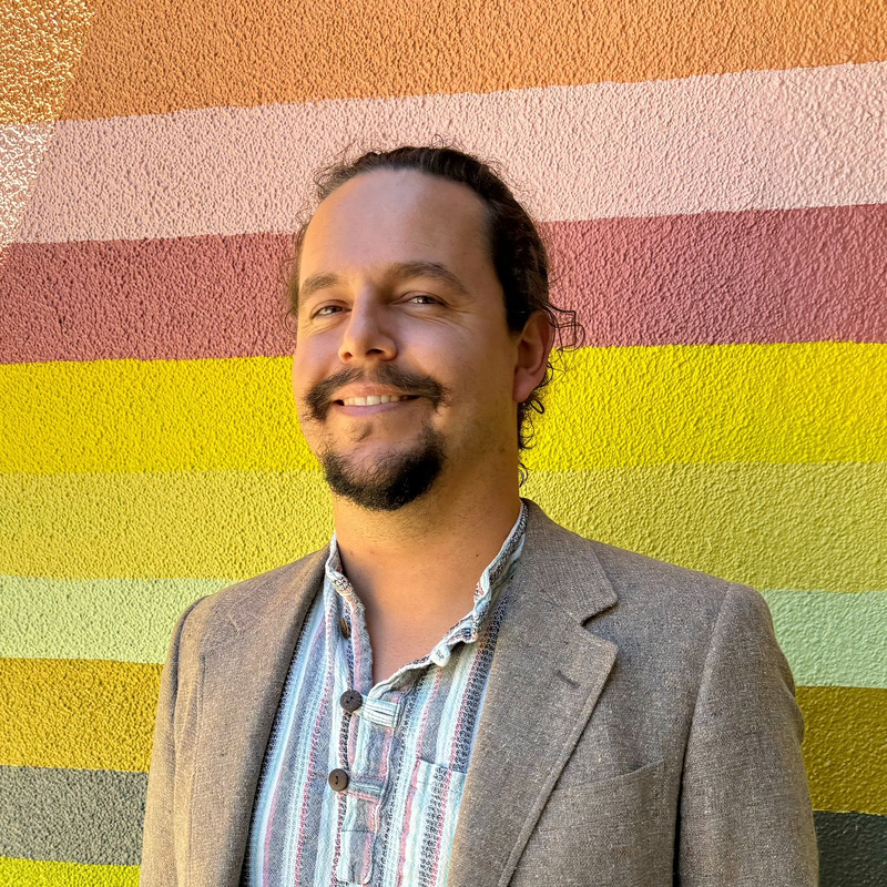 A man with a goatee and jacket stands in front of a rainbow wall