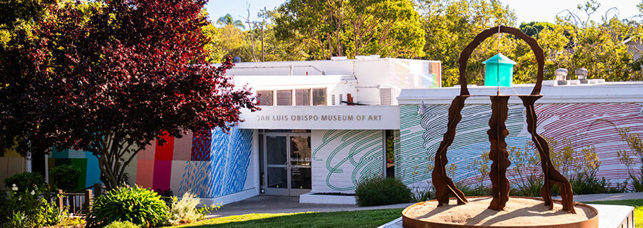 A steel sculpture reminiscent of a jellyfish with a teal resin "house" in the middle. SLOMA in the background with a colorful mural